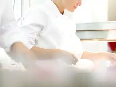 Chef in a white uniform preparing food in a brightly lit kitchen.