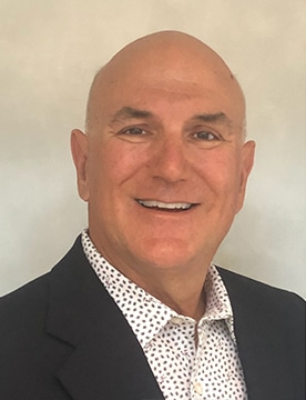 Headshot of Ron Whitfield, board member at Vitalacy, wearing a dark suit and patterned shirt, smiling against a neutral background.